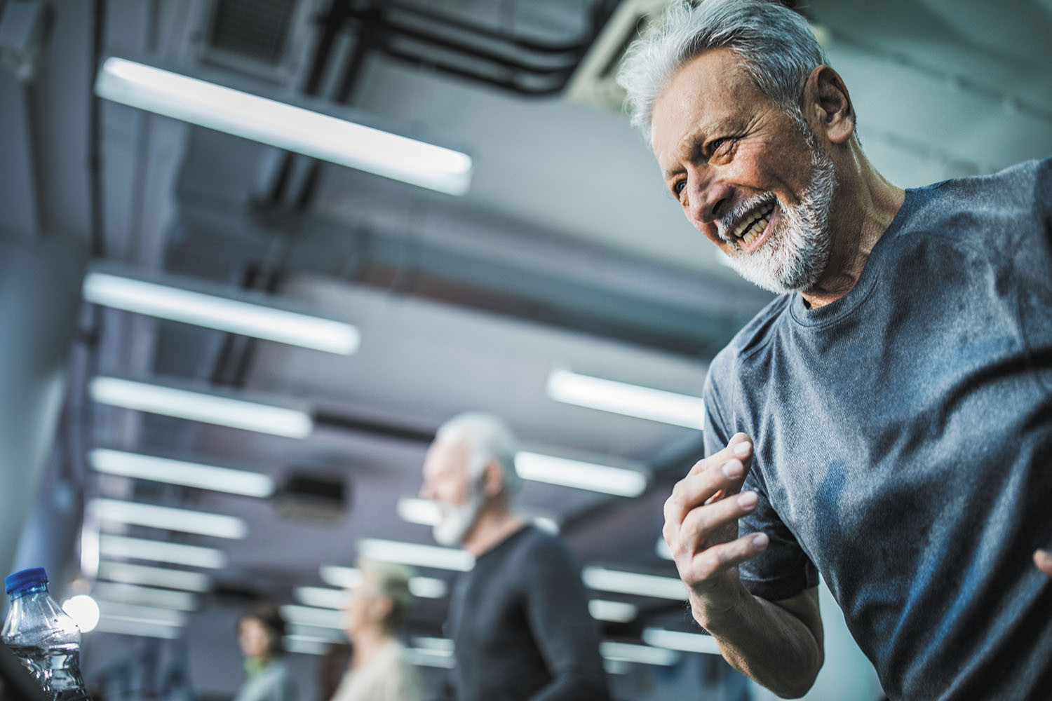 photo of an older man smiling while exercising on a treadmill in a gym; other men are visible behind him